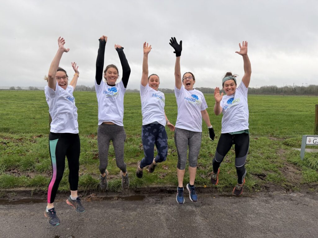 A group of five people wearing Headway Thames Valley T‑shirts jump in unison on a wet path beside a wide grassy field under an overcast sky.